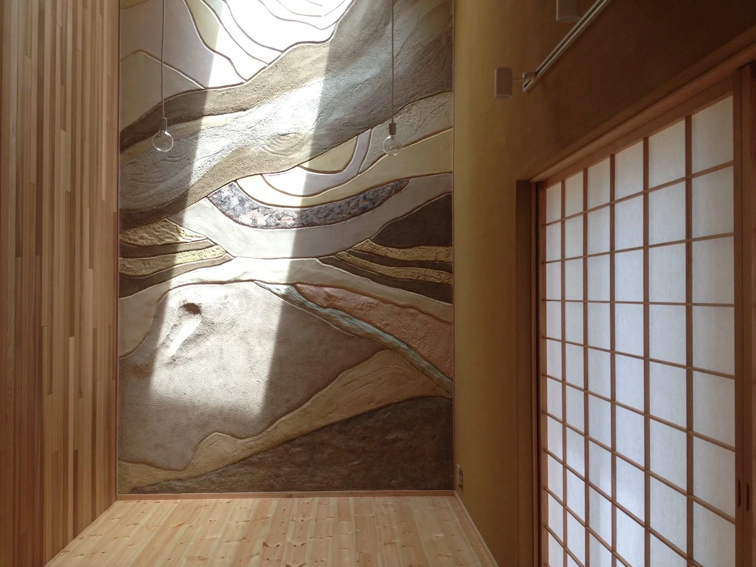 Sculptural earthwork wall with layered organic forms, textured earthen surfaces, and natural light in an interior space. 有機的な層状の造形と土の質感が、自然光によって浮かび上がる室内のアースワーク作品。