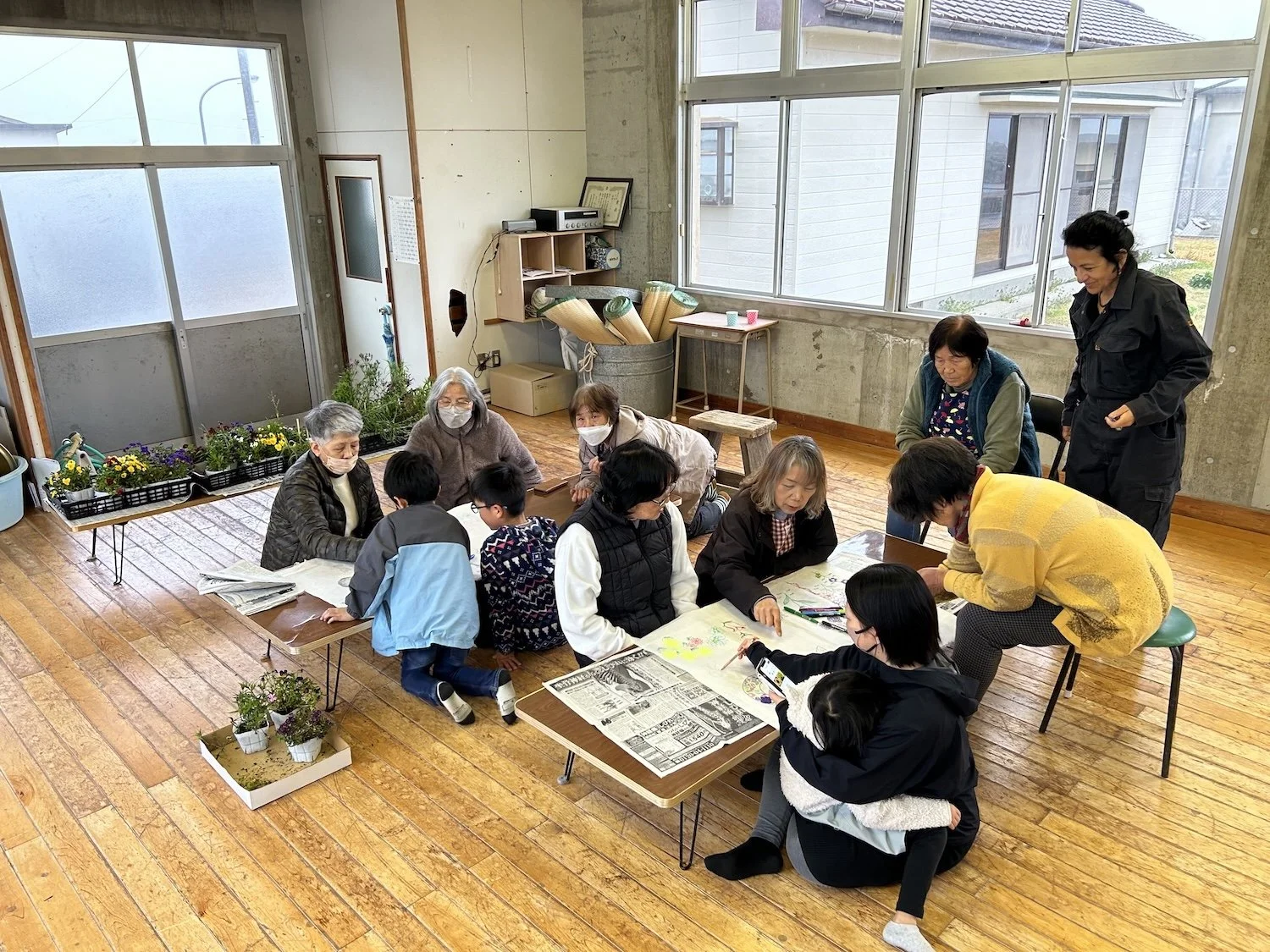 Children and adults gather around a low table making things together during a WAKU WAKU program in Himeshima. 姫島のWAKU WAKUプログラムで、低いテーブルを囲み、一緒に制作する子どもと大人たち。