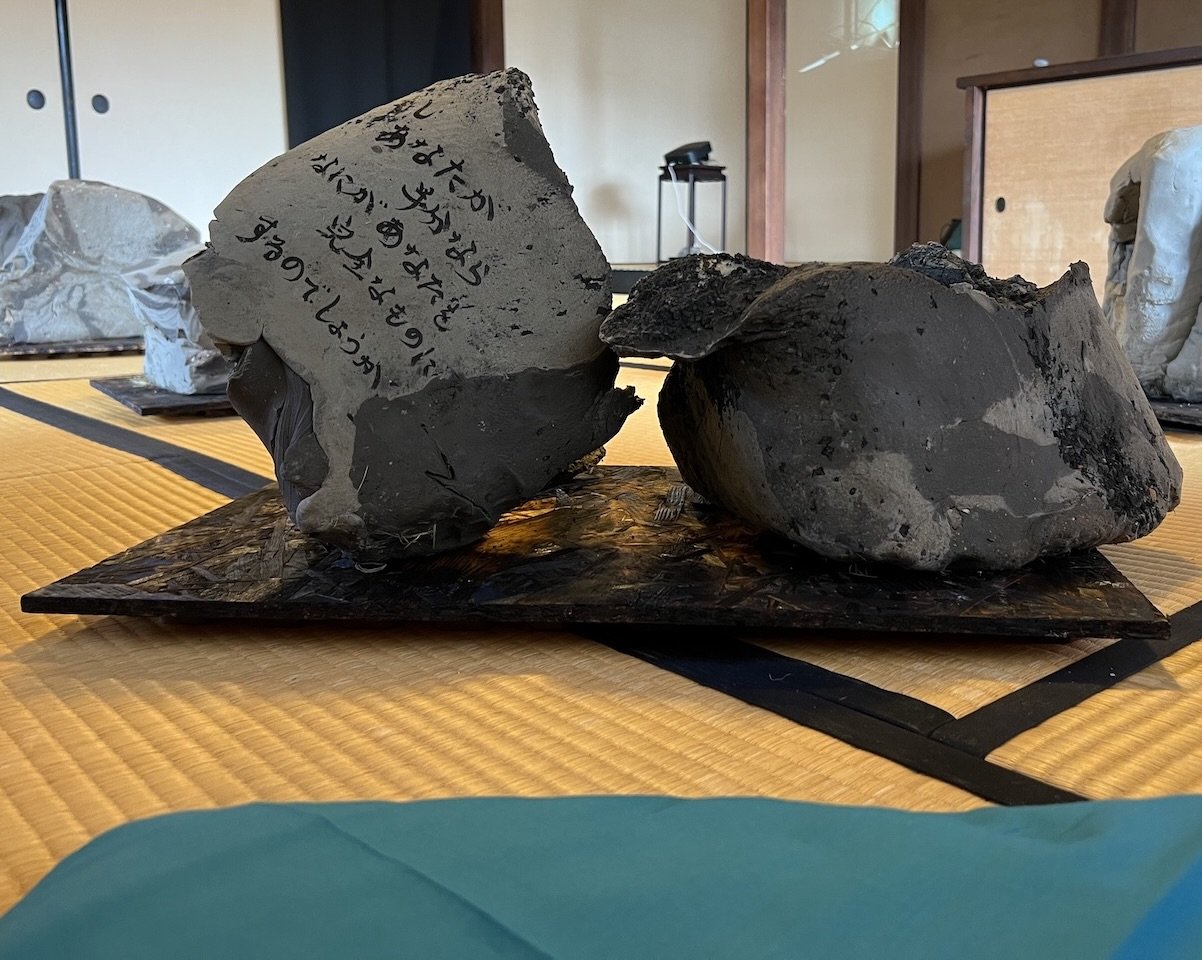 Several irregular raw-clay forms in the Sealed by Time installation, including a central form with handwritten Japanese words, arranged on tatami mats at the Former Nakano Hanroku Residence in Handa, Aichi.