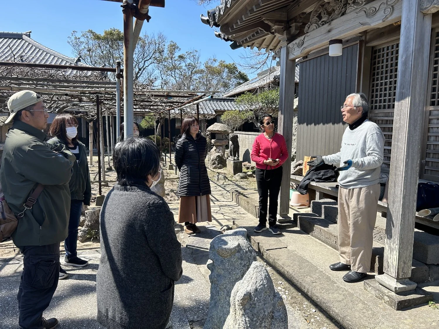 A small group listens to a local speaker outdoors beside traditional buildings in Himeshima. 姫島の伝統的な建物のそばで、屋外で地域の人の話を聞く参加者たち。