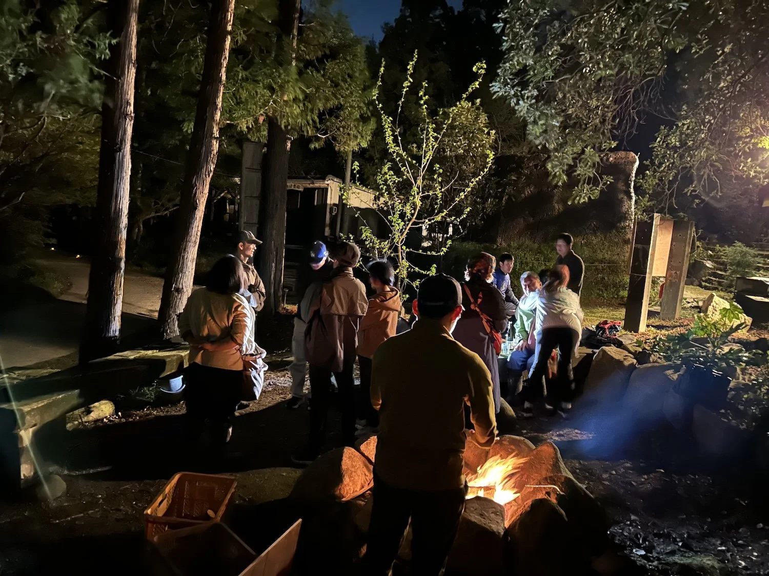 People gather outdoors among trees and warm firelight during an evening 360° Open Hiroba event at Yama no Hiroba. 夜の山の広場®で、木々と火の明かりのそばに集まる360°オープン広場の参加者たち。