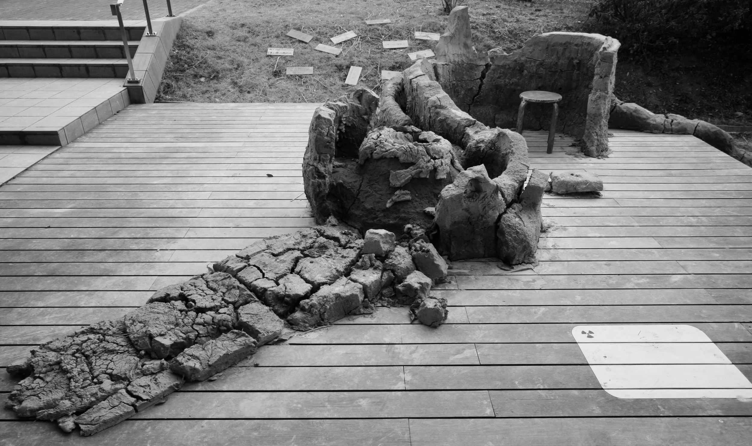 The Common Ground raw-clay installation after exposure outdoors, showing cracked and partially collapsed clay forms beside the wooden deck. 屋外展示を経た《Common Ground》の生土によるインスタレーション。木製デッキのそばで、土の形がひび割れ、一部が崩れている様子。
