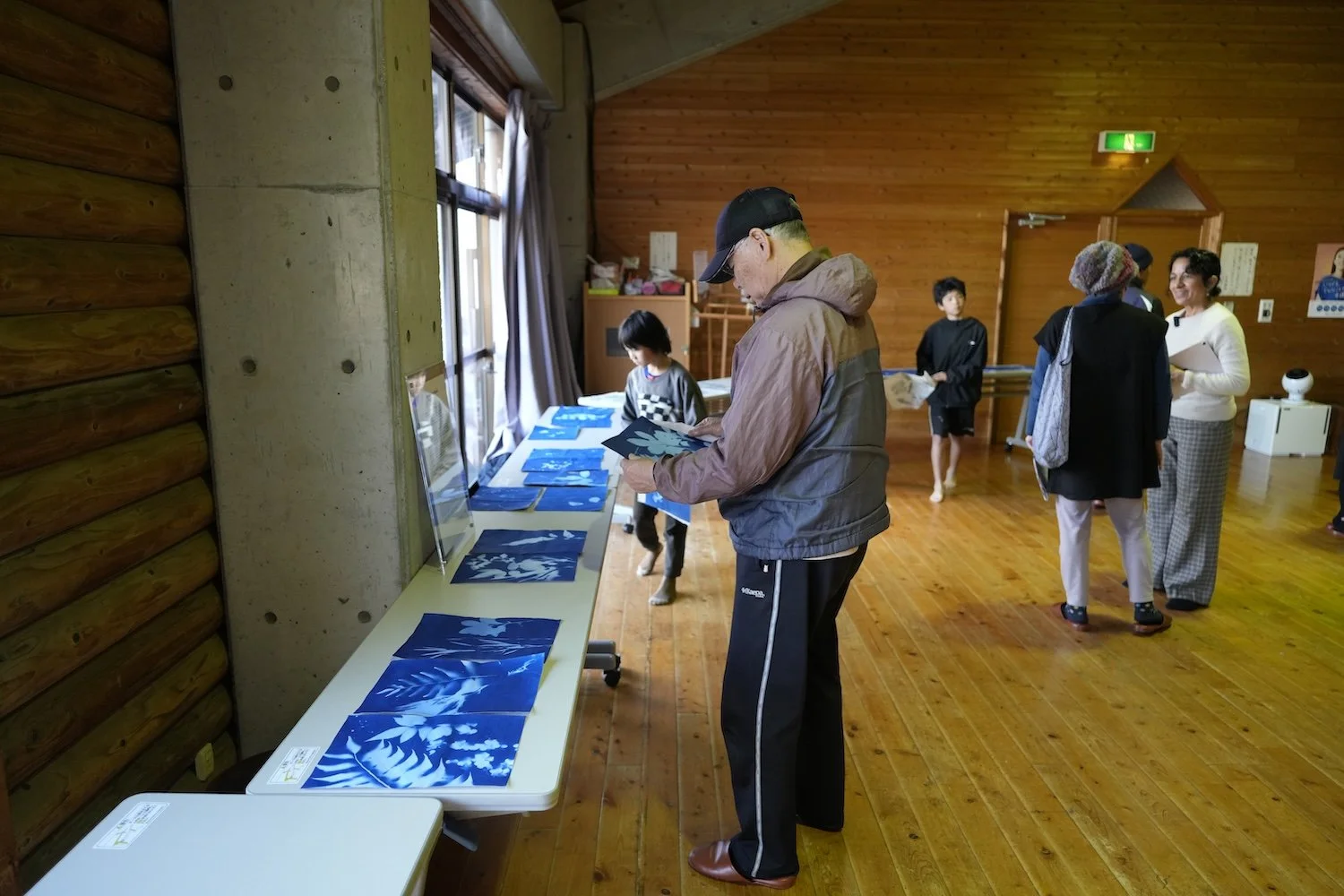 Visitors view cyanotype prints and other GEO–OASIS documentation displayed on tables during the public exhibition in Himeshima. 姫島で行われたGEO–OASISの展示で、テーブルに並べられたサイアノタイプ作品や記録を見る来場者。
