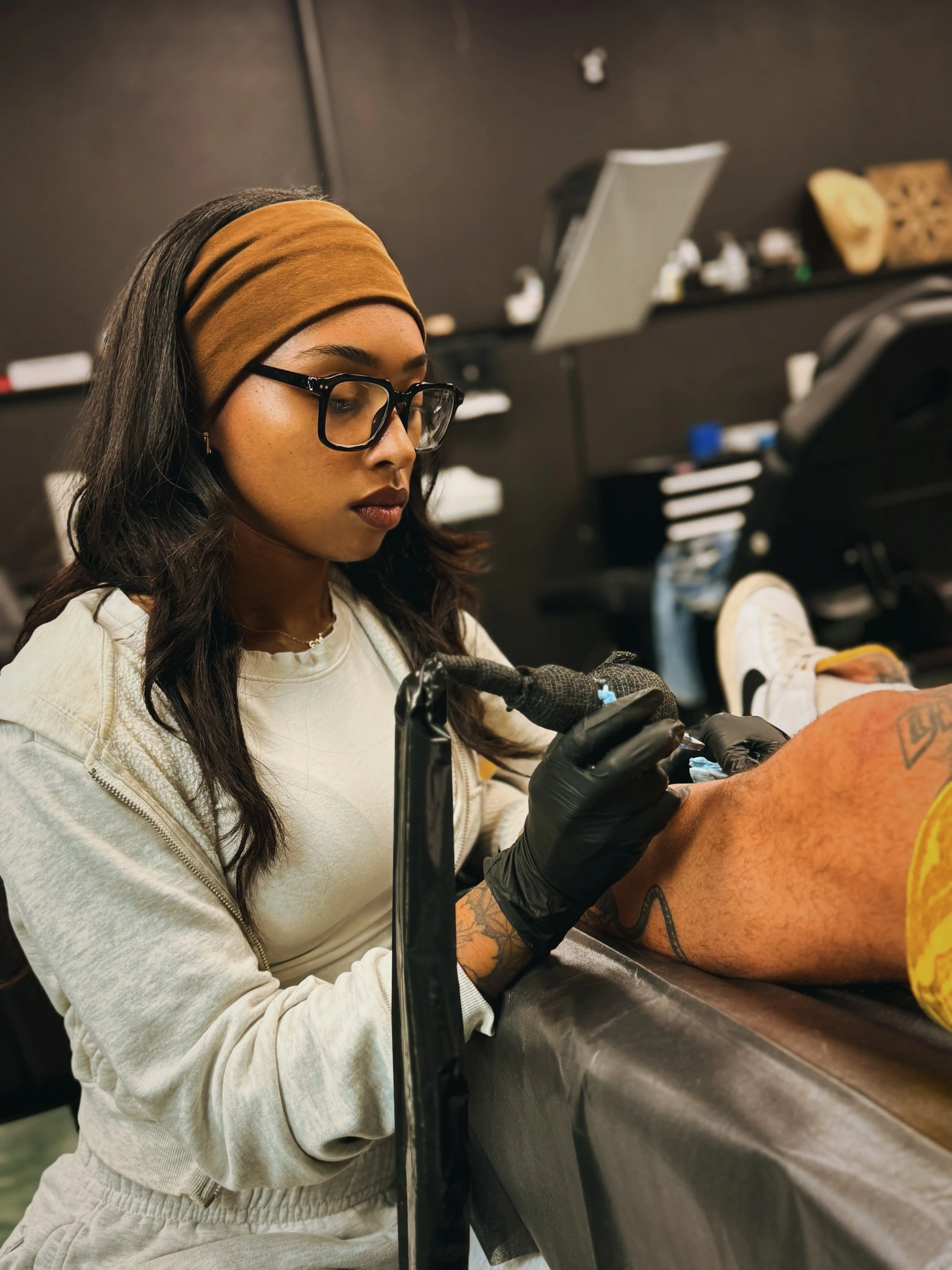 A woman with glasses and a brown headband is tattooing a person's leg in a tattoo parlor.