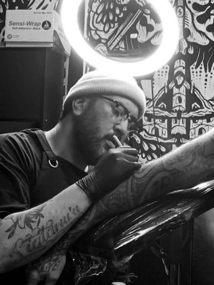 A man with tattoos on his arm, wearing glasses, a beanie, and gloves, is sitting at a tattooing station. He is holding a tattoo machine and appears to be tattooing. There is artwork on the wall behind him, and a bright circular light is above his head.