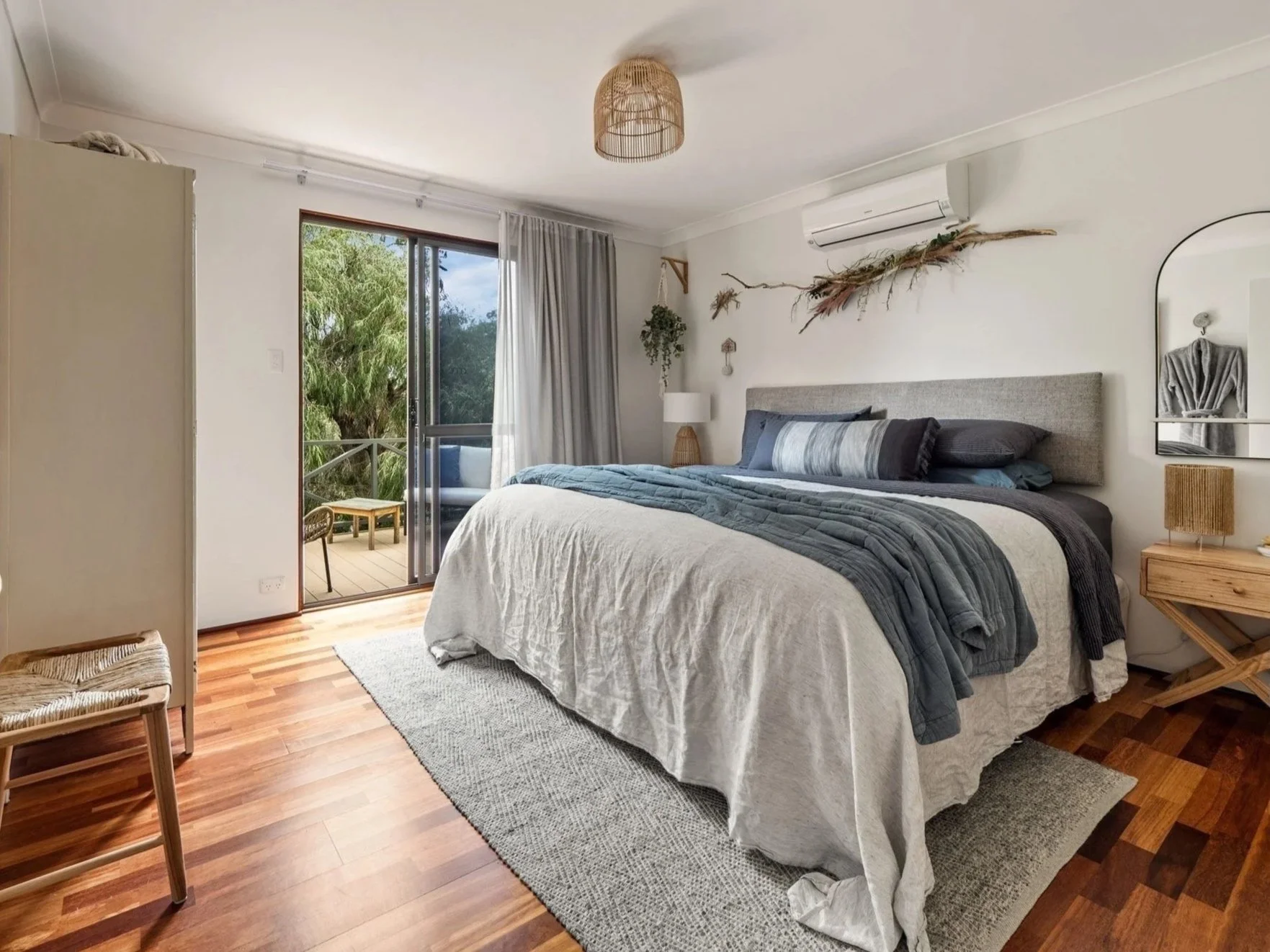 Modern bedroom with a large bed, blue and gray bedding, wooden flooring, and a sliding glass door leading to a balcony. There is a wall-mounted air conditioner, small side table, lamp, and decorative elements like plants and artwork.