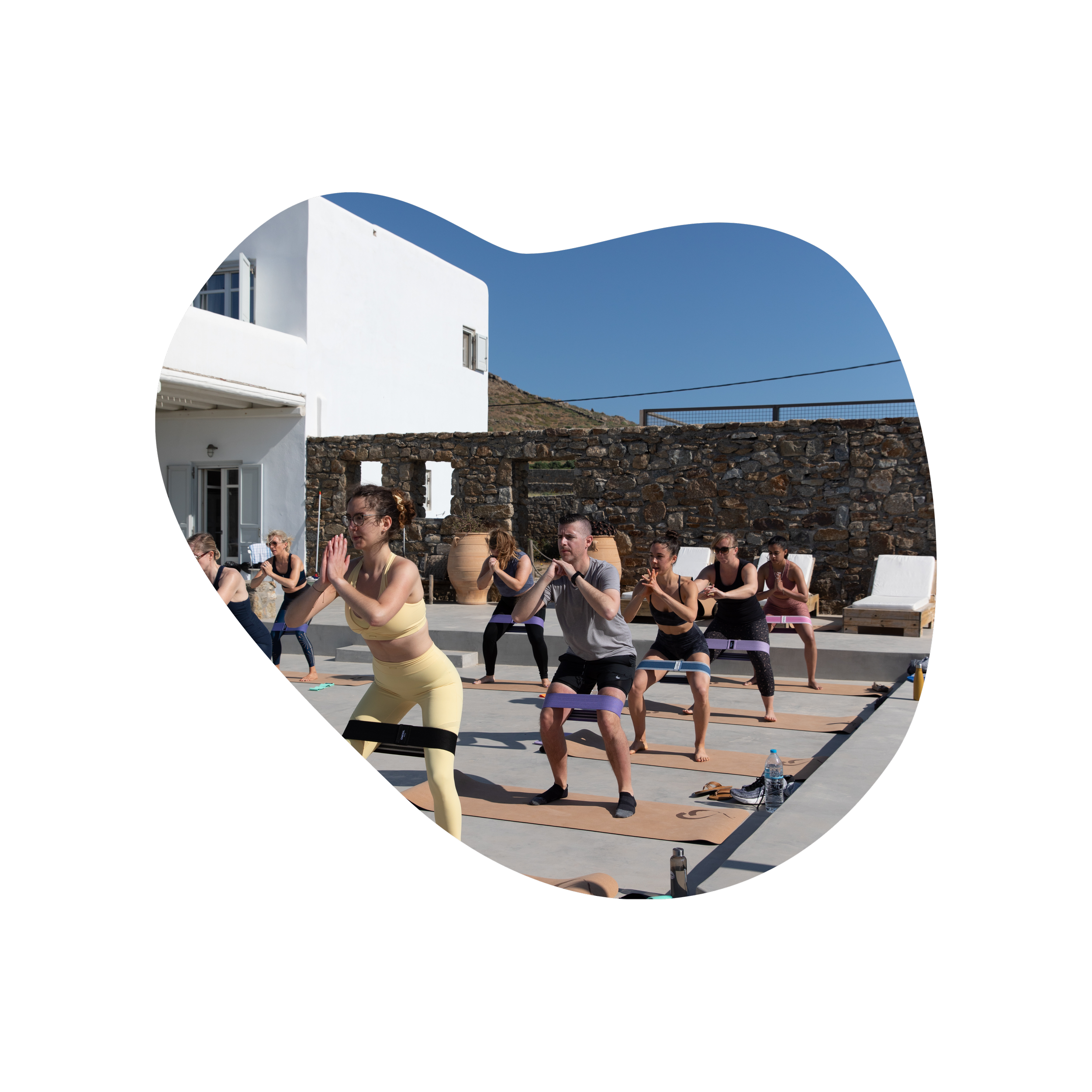People participating in outdoor yoga class on a rooftop patio with a stone wall, white buildings, and clear blue sky in the background.