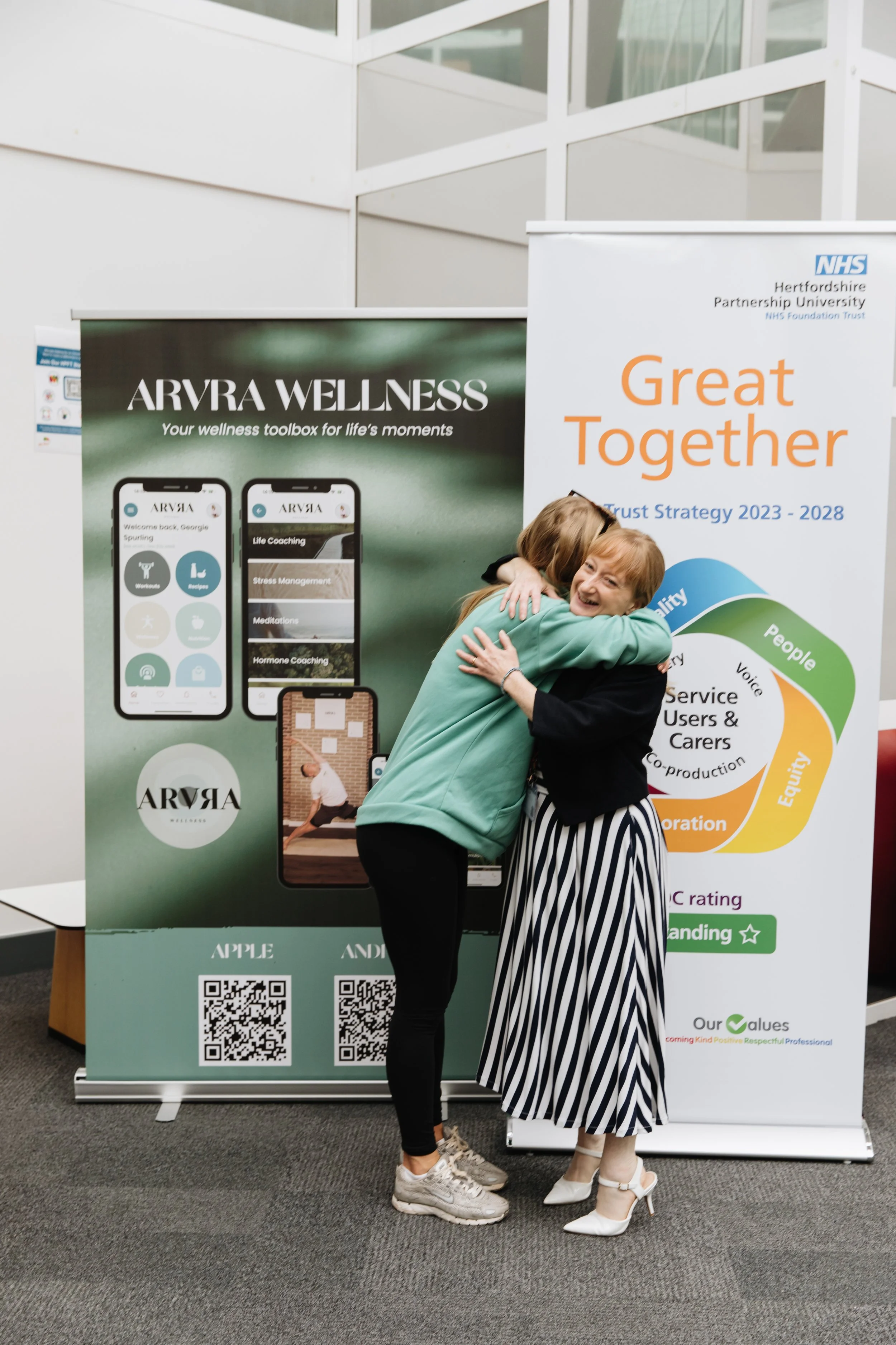 Two women hugging and smiling at an indoor event with wellness and partnership banners in the background.