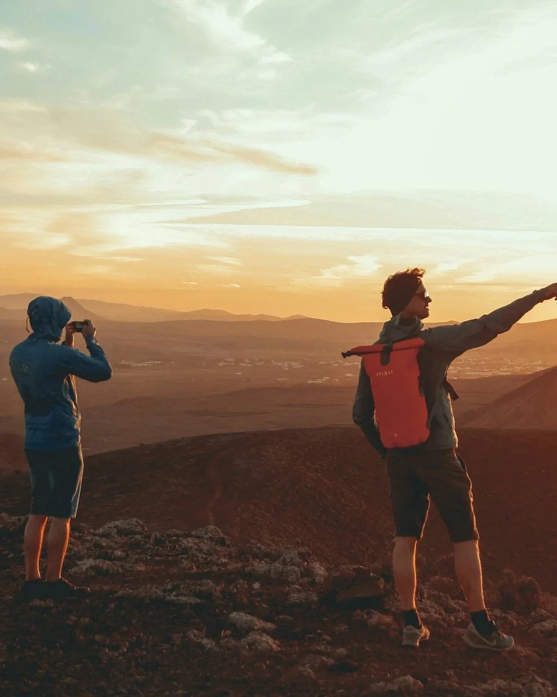 Two hikers standing on a rocky terrain during sunset, one taking a photo with a camera, the other pointing towards the horizon, with mountains and a valley in the background.