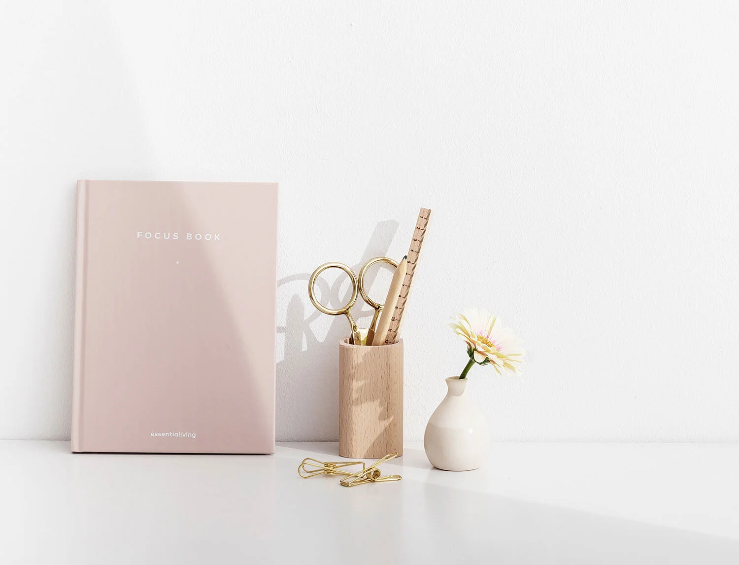 A workspace with a pink notebook labeled 'Focus Book', gold scissors, a wooden ruler, a small vase with a flower, and some gold paper clips on a white surface against a white wall.