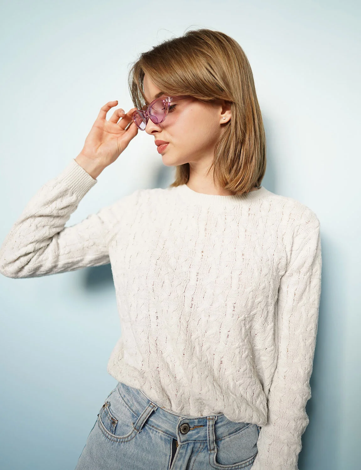 A young woman with shoulder-length light brown hair is wearing pink sunglasses and a white cable-knit sweater. She is standing against a light blue background, looking down and touching her sunglasses with her right hand.