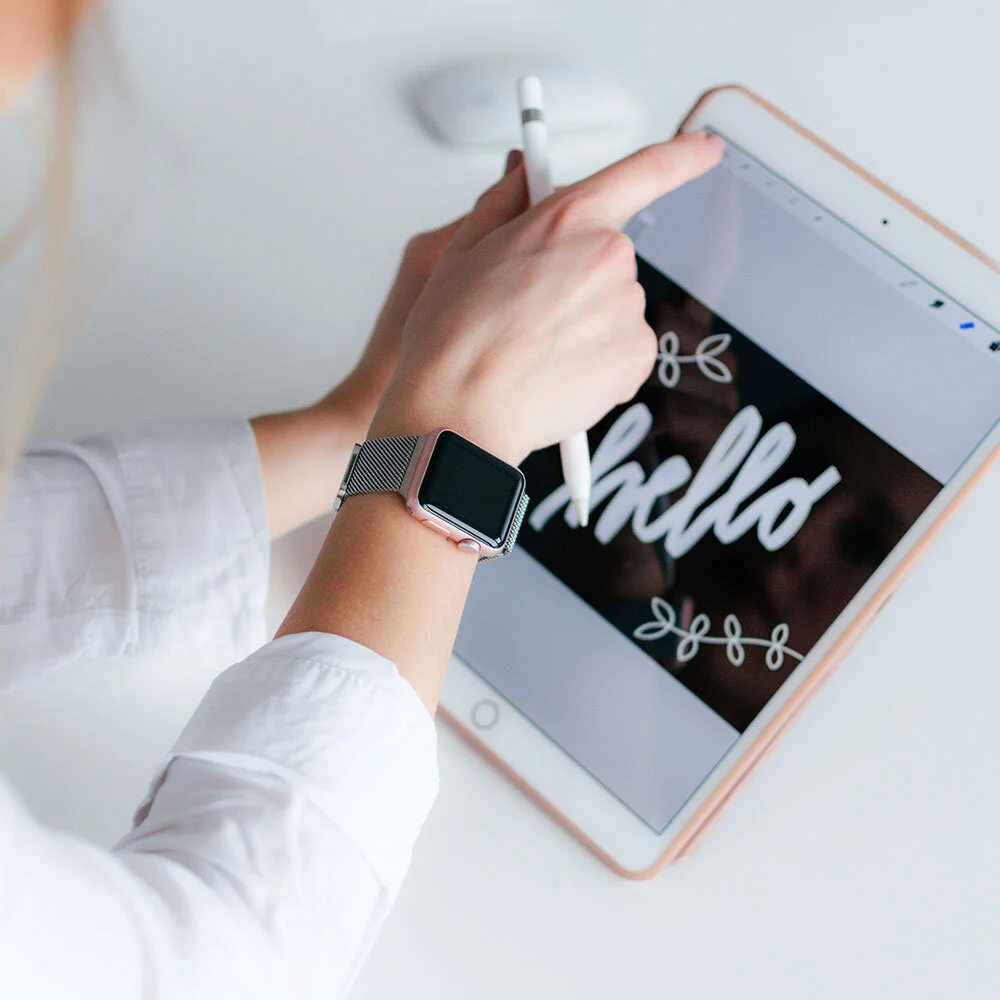 Person using an iPad and Apple Pencil, wearing a smartwatch, with a white shirt, on a white surface, displaying the word 'Hello' in a handwritten style.