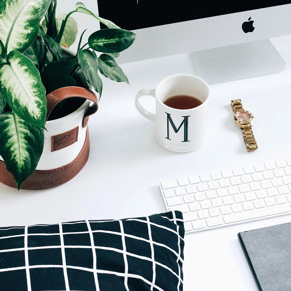 A desk with a potted plant, a mug with the letter M, a gold wristwatch, a white keyboard, an iMac computer, a closed notebook, and a black-and-white checkered pillow.