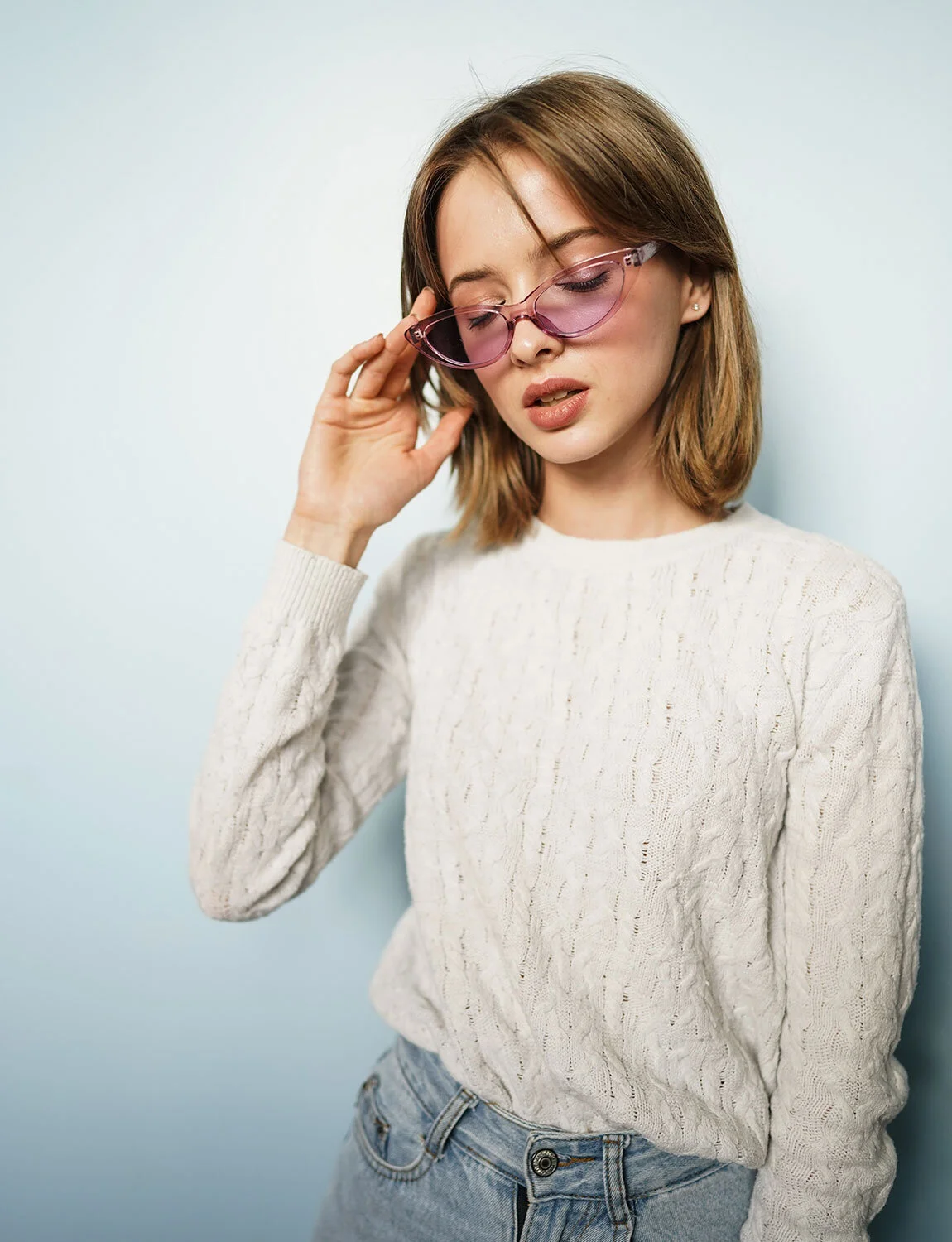 A woman with shoulder-length brown hair wearing pink sunglasses, a cream knit sweater, and blue jeans, standing against a blue background.