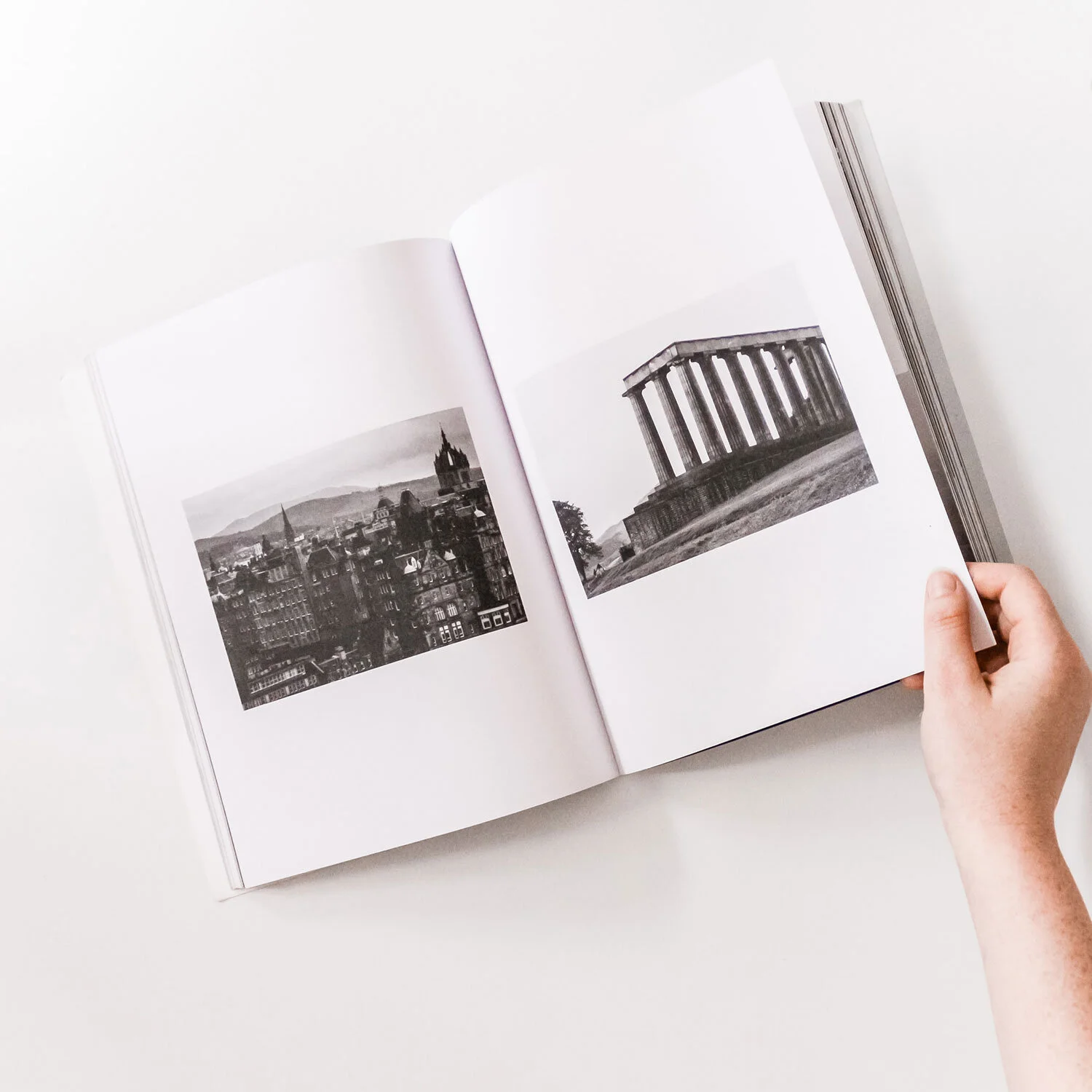 Open book with black and white photos of a cityscape and ancient Greek temple ruins on white pages, held by a person's hand on a white surface.