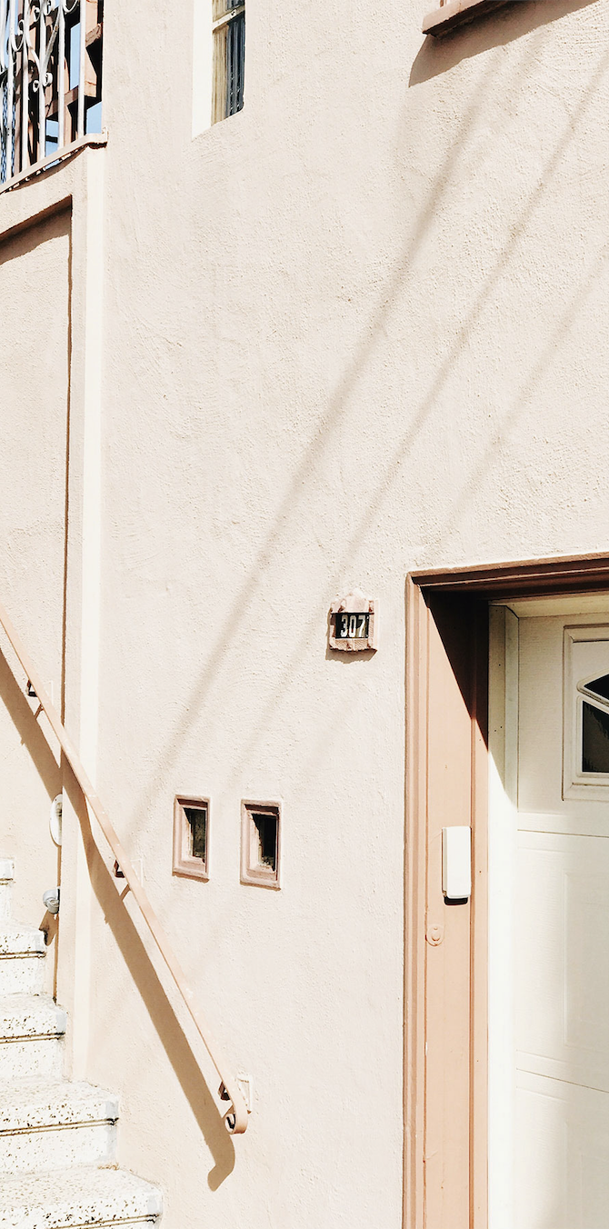 Exterior of a beige stucco house with small decorative windows, a staircase with a metal railing, and a garage door. The house number is 307, mounted on a small brick plaque.