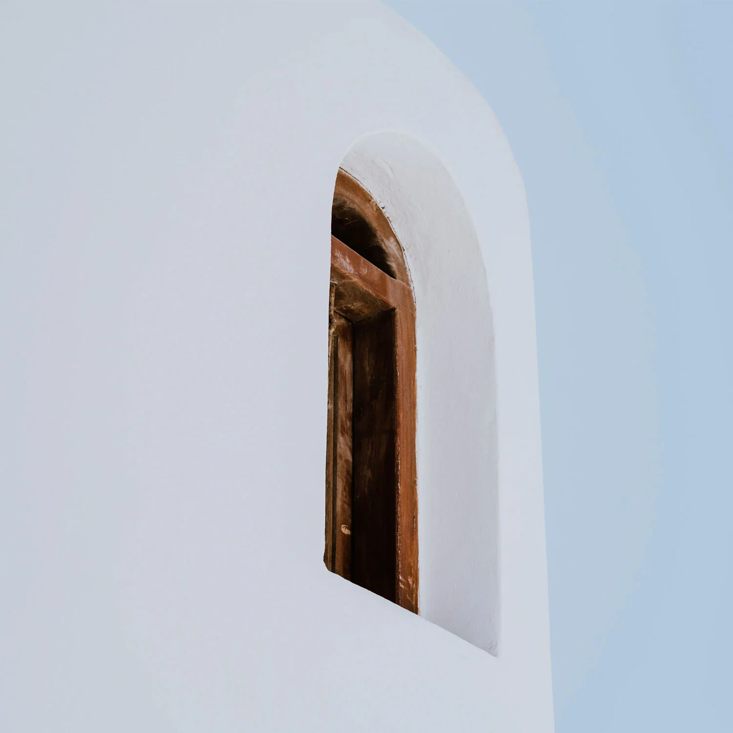 A small arched window with a wooden shutter on a white wall, with a light blue sky in the background.