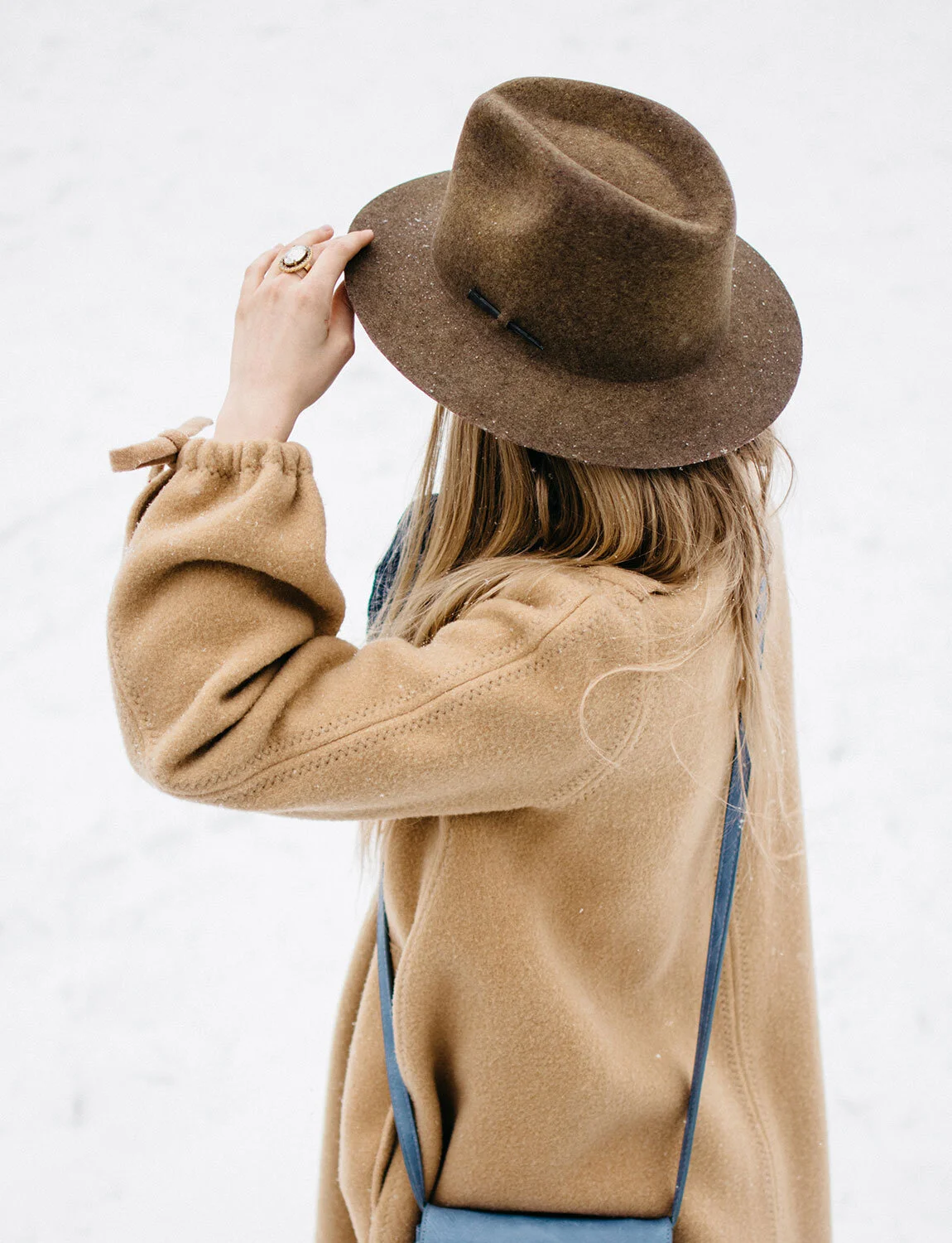 A person wearing a tan coat and a brown hat, with long blonde hair, standing against a snowy background and holding the brim of their hat.