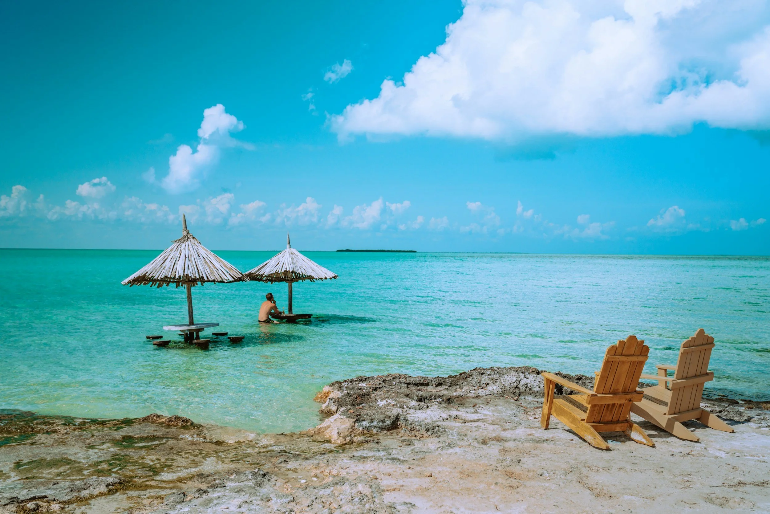 Beach scene with two wooden Adirondack chairs on the sandy shore and two thatched umbrellas over a picnic table and a person sitting in the ocean.