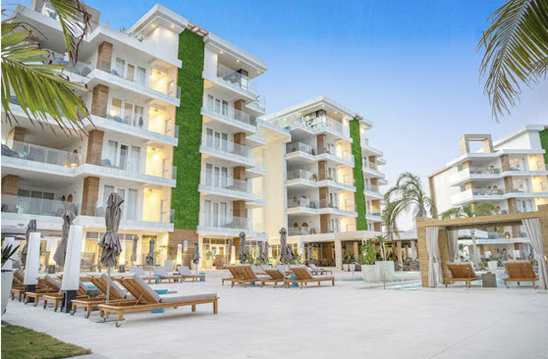 A modern hotel with multiple stories, outdoor pool area, and lounge chairs, surrounded by palm trees during dusk.