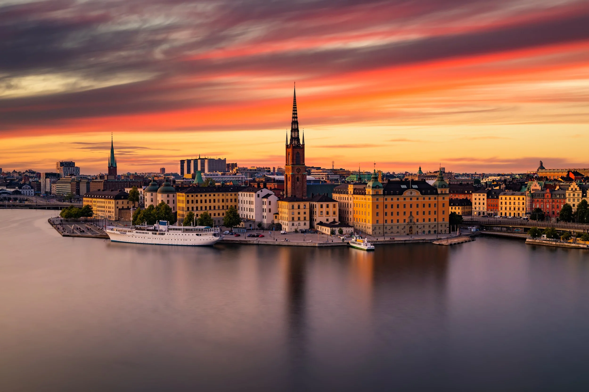 Sunset over a city with historic buildings and a body of water reflecting the skyline, including a tall church with a spire and several boats docked along the waterfront.