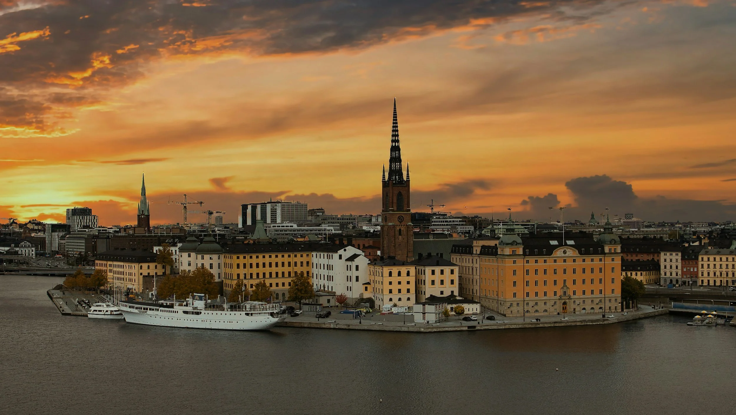 View of a city skyline at sunset with historic buildings, a river with a white ship docked along the shore, and a dramatic orange and yellow sky with clouds.