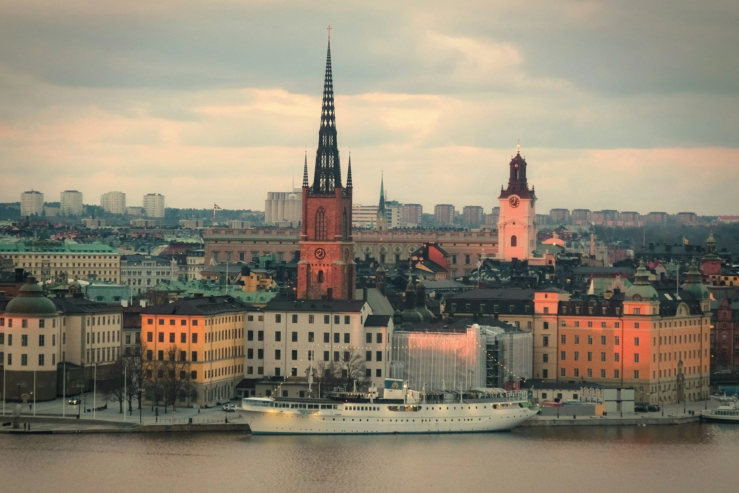 Cityscape of Stockholm, Sweden, with church steeples, historic buildings, and a boat on the water at sunset.