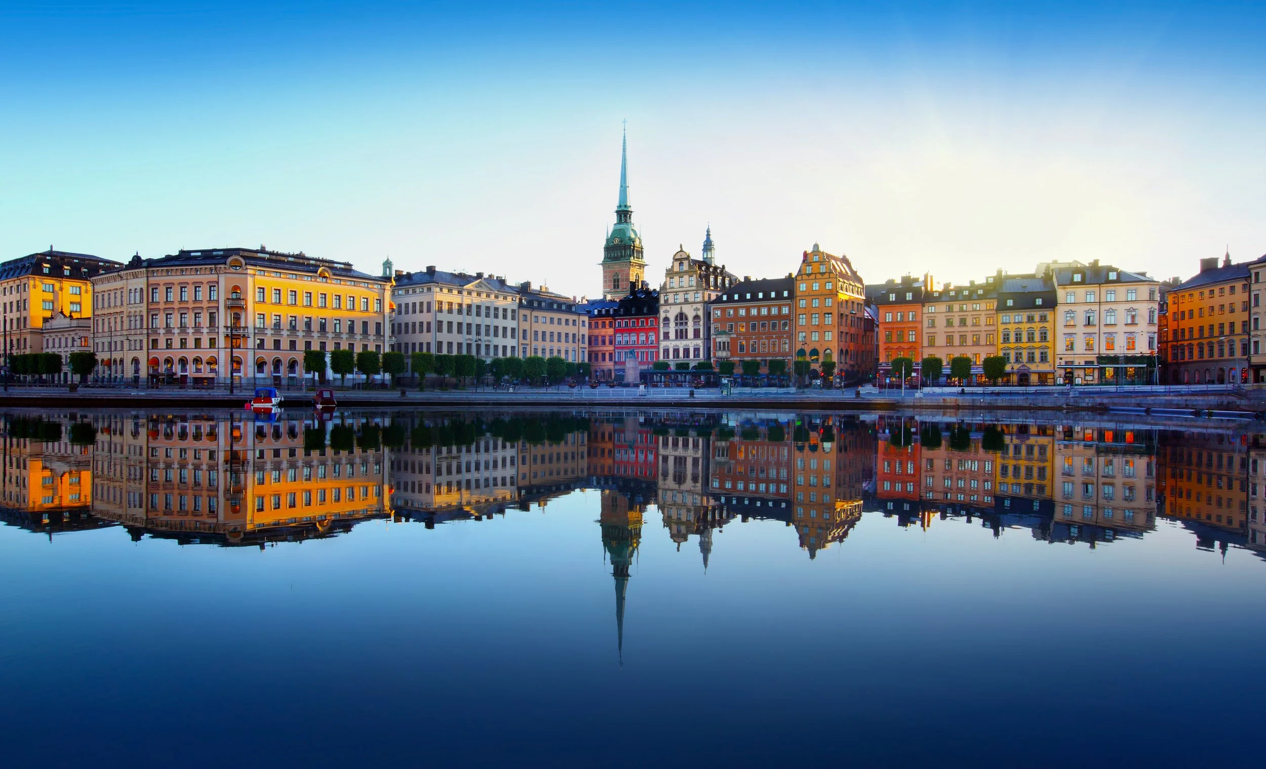Colorful buildings along a river with their reflection visible in the water, under a clear blue sky.