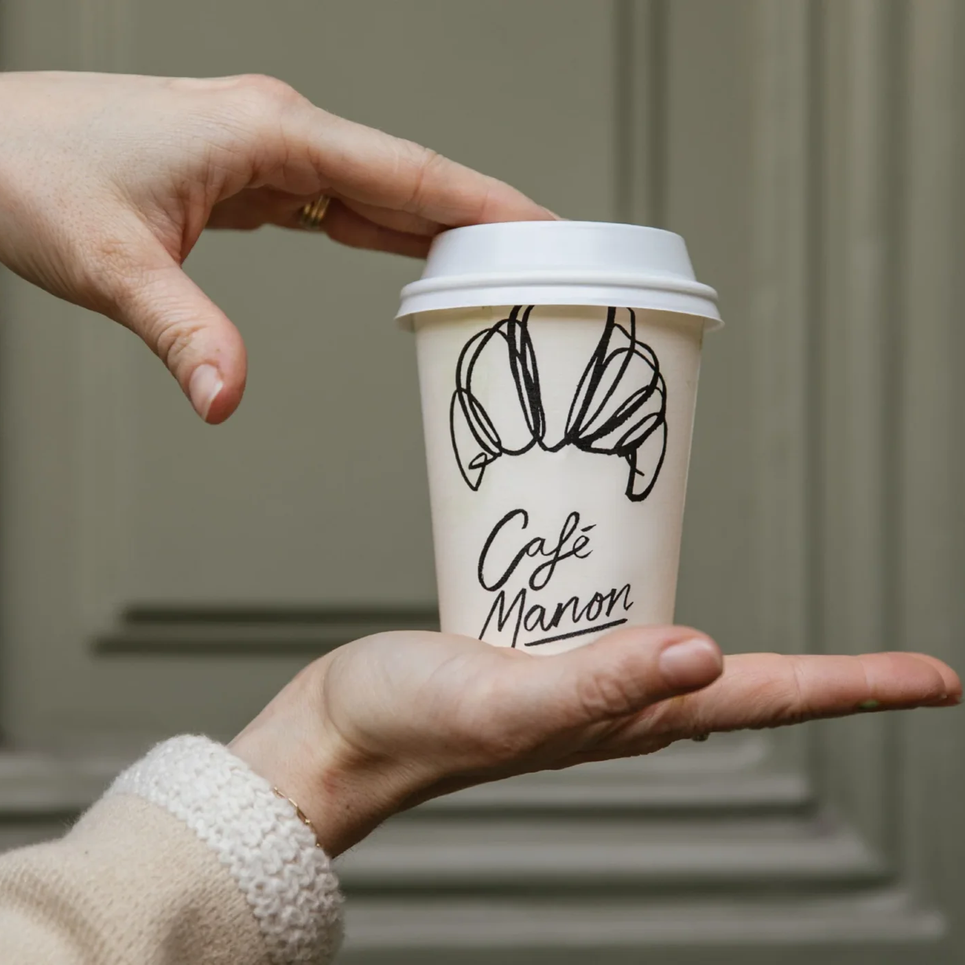 Hands holding a takeaway coffee cup from Café Manon featuring a croissant illustration and handwritten logo, symbolising artisan coffee, pastries and a cosy Parisian-style café experience.