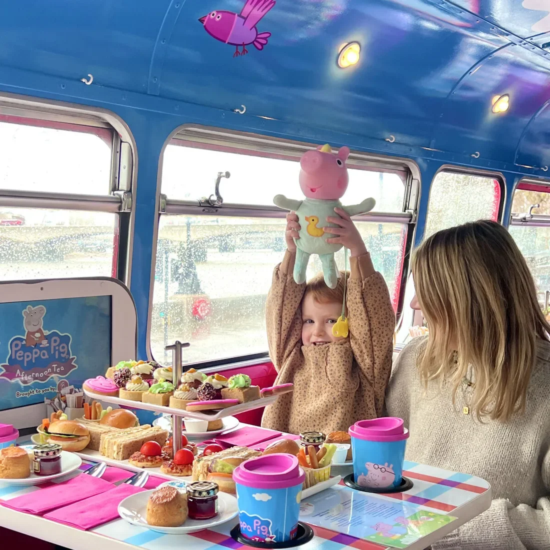 Child holding Peppa Pig toy on a themed bus while enjoying afternoon tea with parent, surrounded by colourful snacks and decorations