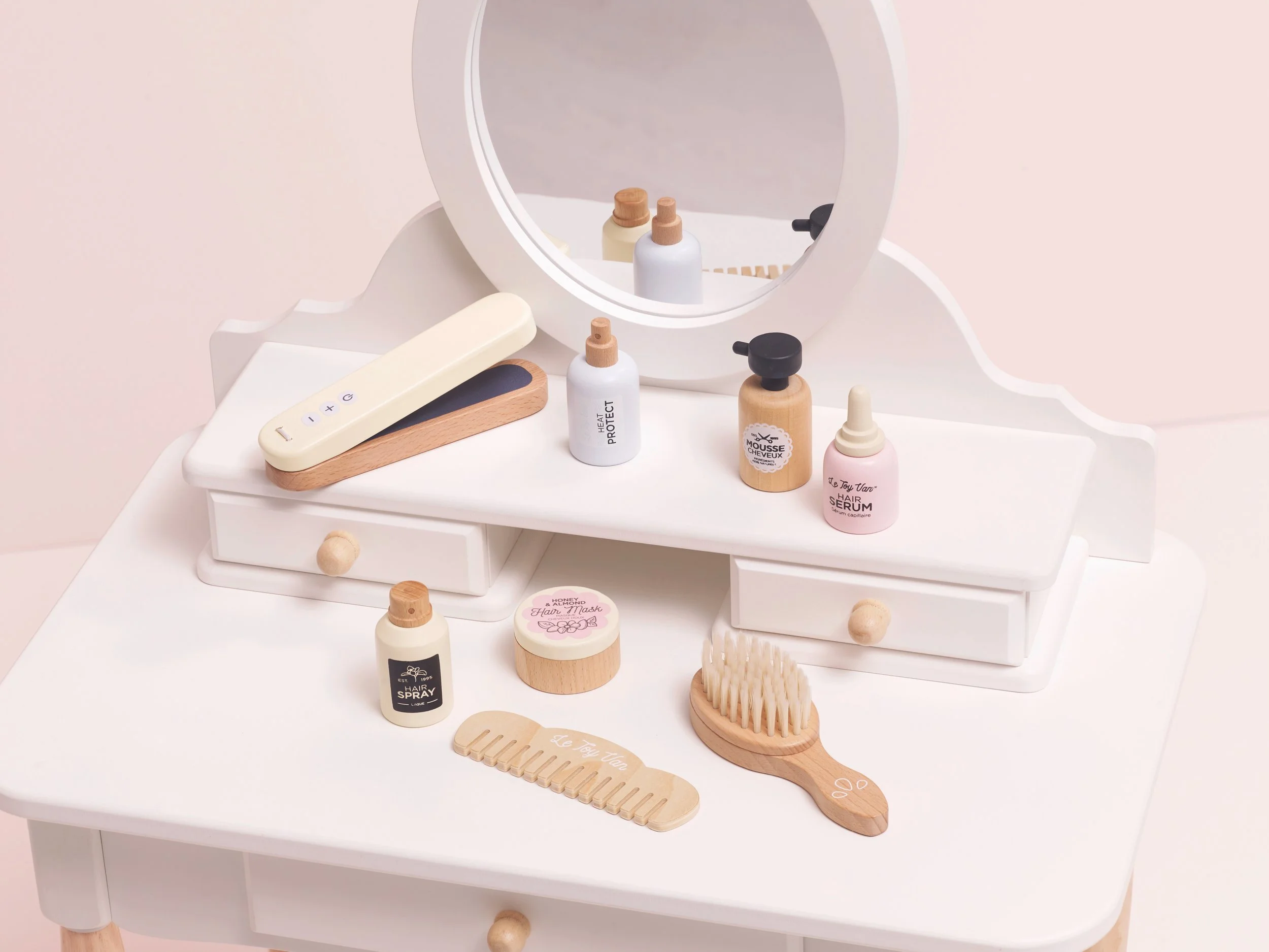 A white toy vanity table with a round mirror displays miniature wooden beauty accessories, including brushes, a comb, pretend bottles, and nail files arranged neatly on the surface.