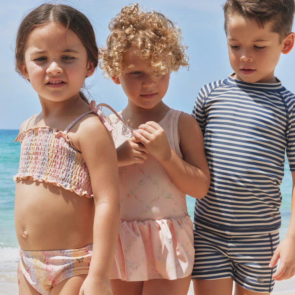 Three children wearing summer swimwear stand by the sea, including a patterned bikini, a floral swimsuit and a striped short-sleeve swim set.
