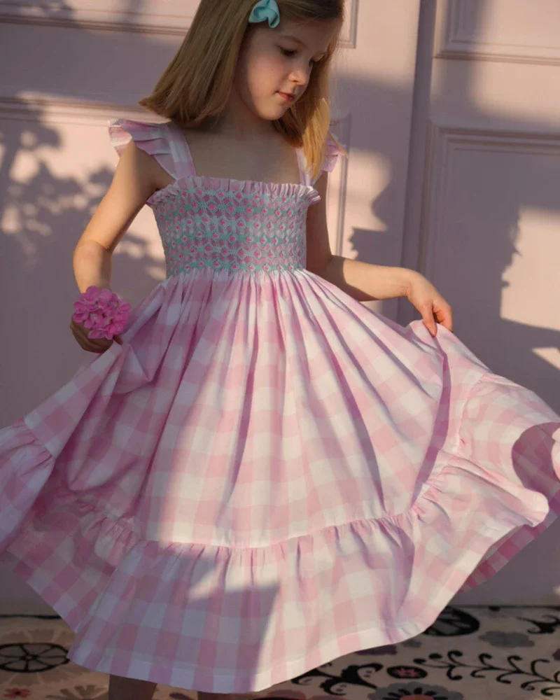 Girl wearing pink gingham smocked dress with ruffle straps, lightweight spring summer children’s outfit in soft natural light
