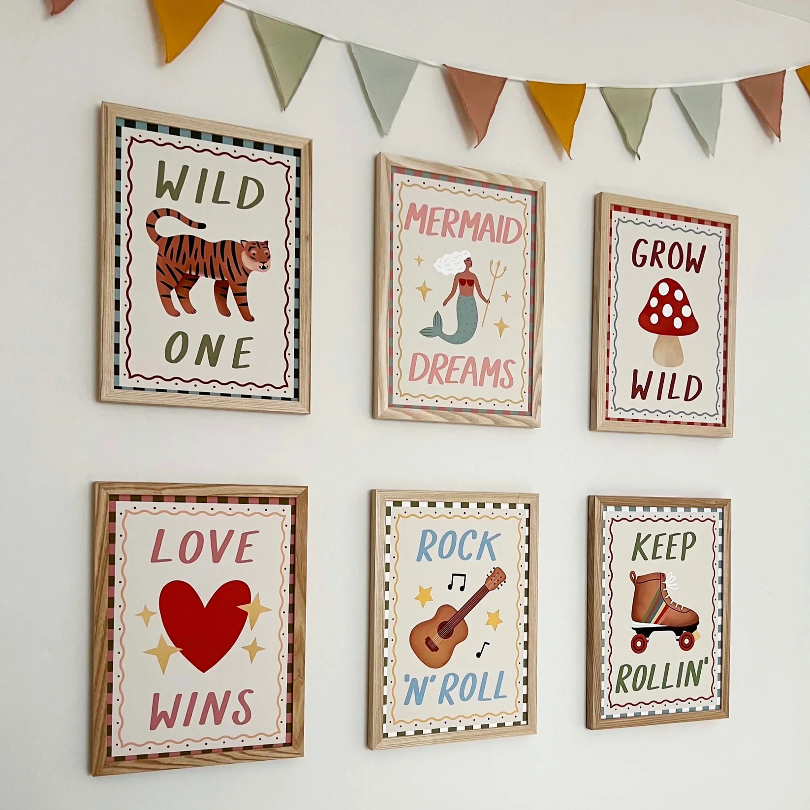 Gallery wall of six framed children’s art prints with playful illustrations and phrases, including a tiger, mermaid, heart, guitar and roller skate, displayed beneath pastel bunting.