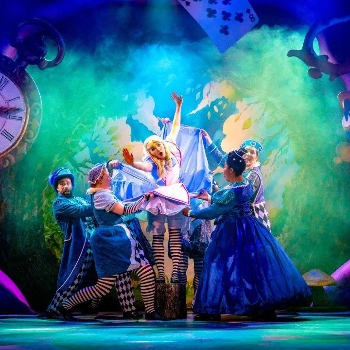 Colourful stage scene from Alice in Wonderland Live featuring Alice and the ensemble cast performing in a magical Wonderland setting with oversized clocks, vibrant costumes and theatrical lighting in a family theatre production.