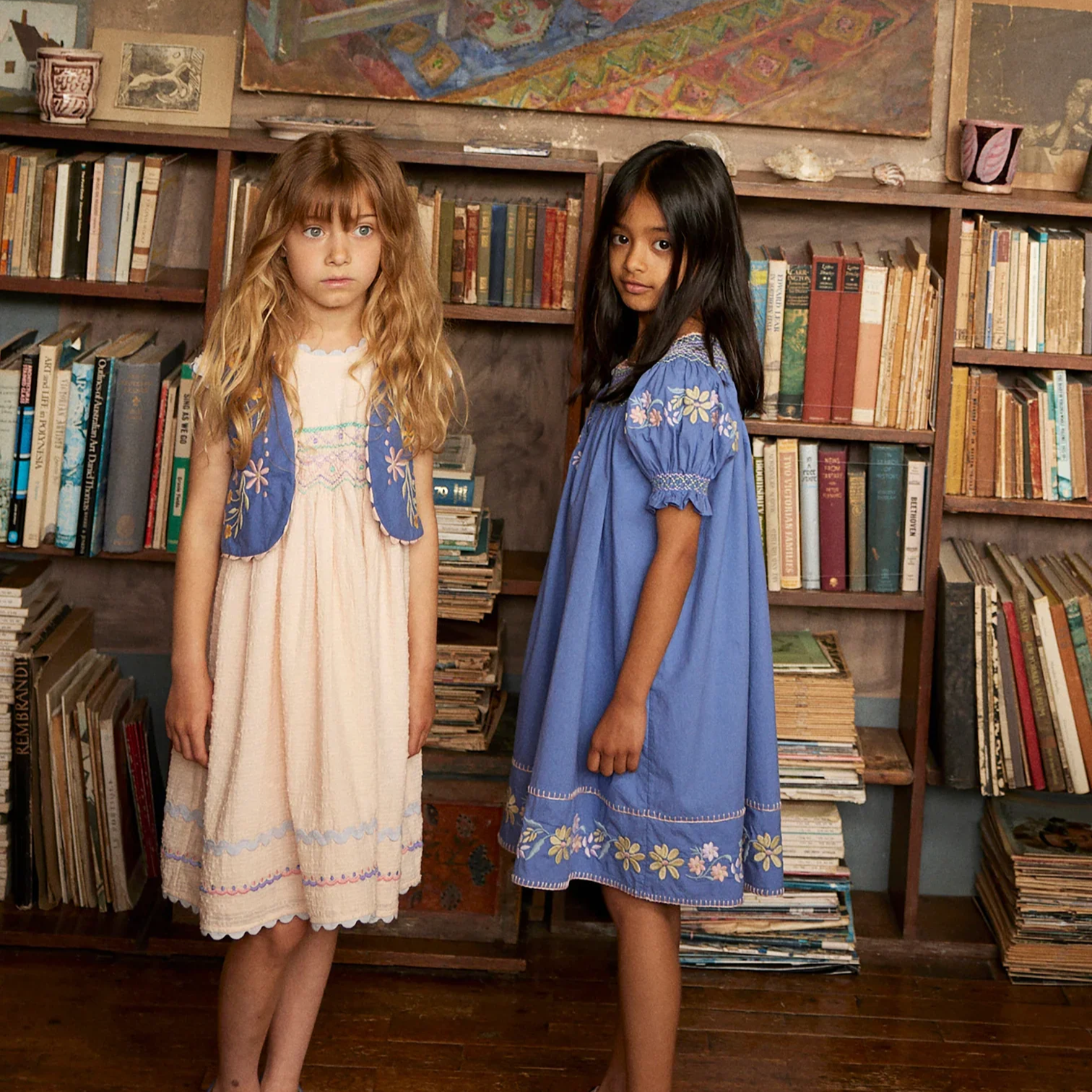 Two girls stand in front of a vintage bookshelf and artwork wearing Apolina Kids dresses, including the Verna Smock Dress in Azul, featuring delicate floral embroidery and soft cotton fabric in a timeless, heritage-inspired style.