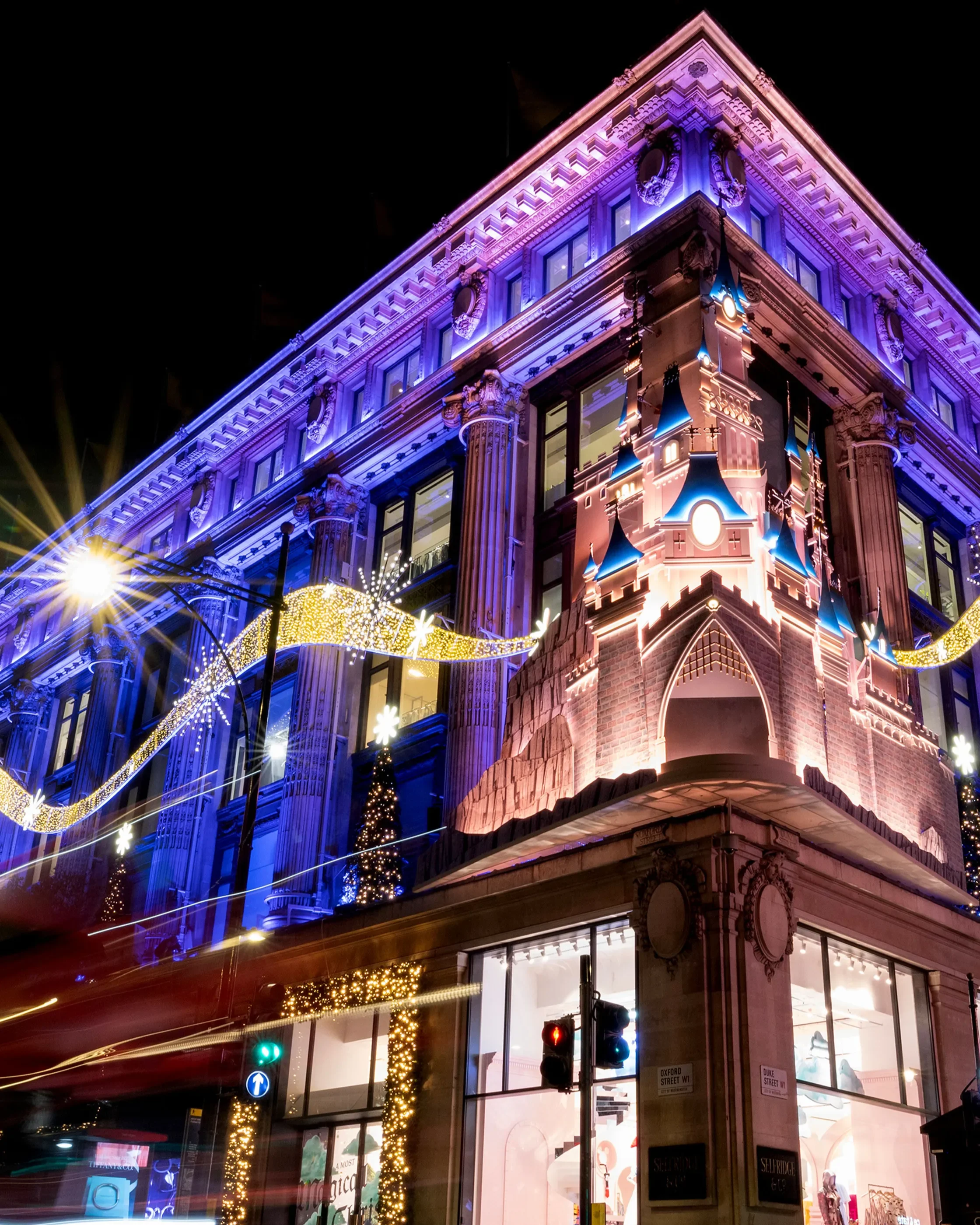 The exterior of Selfridges London lit up at night with vibrant purple and blue Christmas lights, featuring a large illuminated Disney-style castle façade and sparkling festive decorations along the building.
