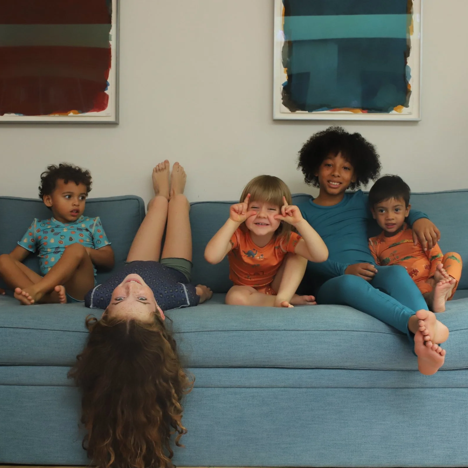 Five children in colourful pyjamas relax and play on a light blue sofa — one lying upside down, one making a peace-sign pose, and the others sitting and smiling together.