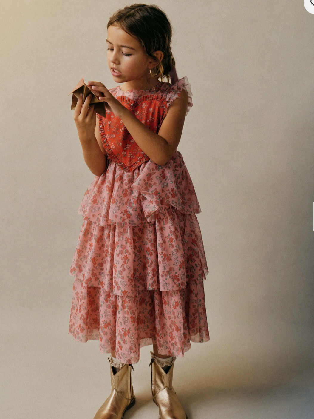 Child wearing a pink floral tiered dress with ruffled sleeves and gold ankle boots, holding a small paper object against a neutral background.