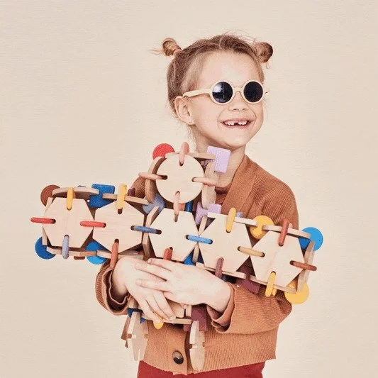 A smiling young child wearing round sunglasses holds a large colourful wooden construction made of interlocking shapes. The child is dressed in a brown cardigan and stands against a neutral background.