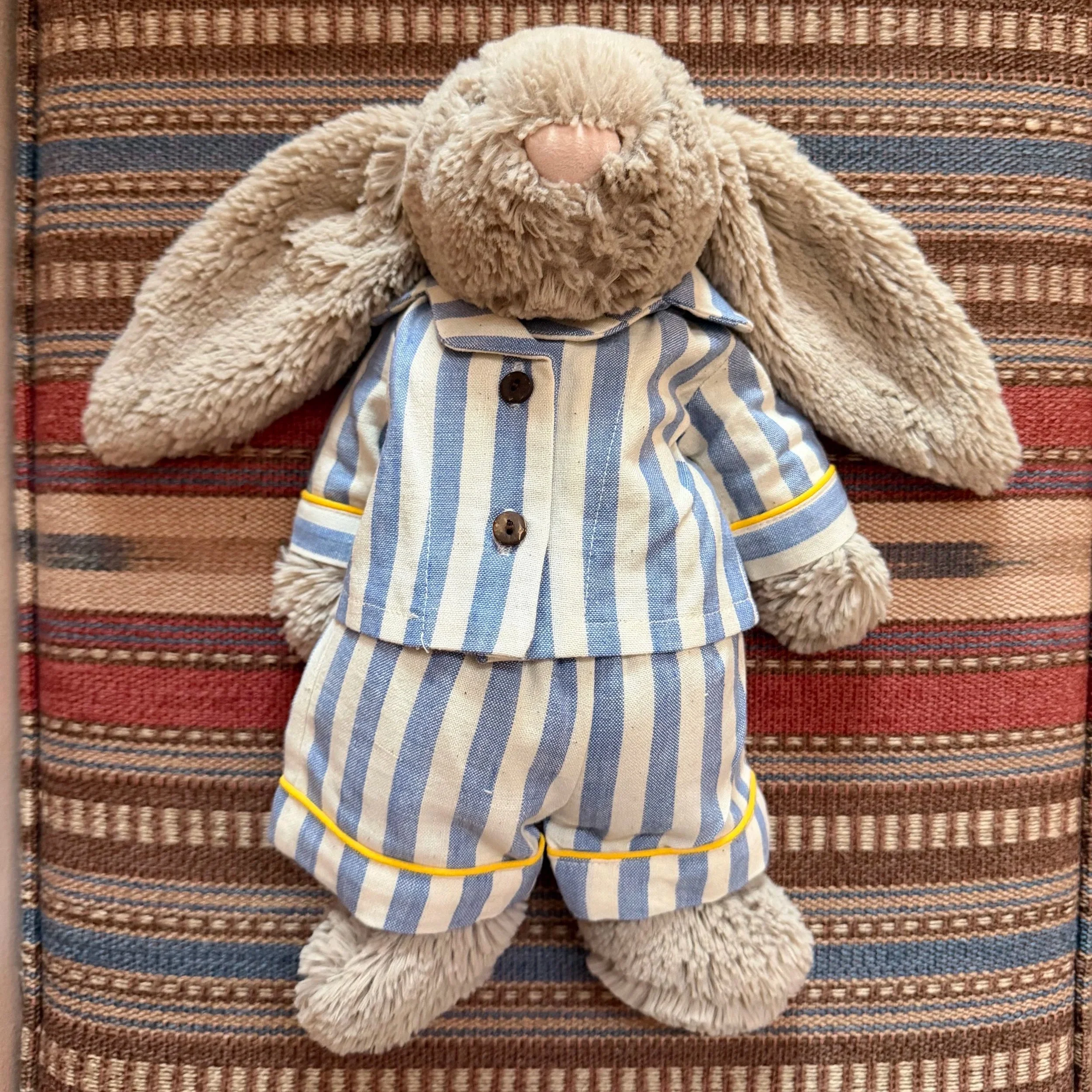 A soft grey cuddly bunny lies on a patterned fabric surface, dressed in blue-and-white striped pyjamas with yellow trim and wooden buttons.
