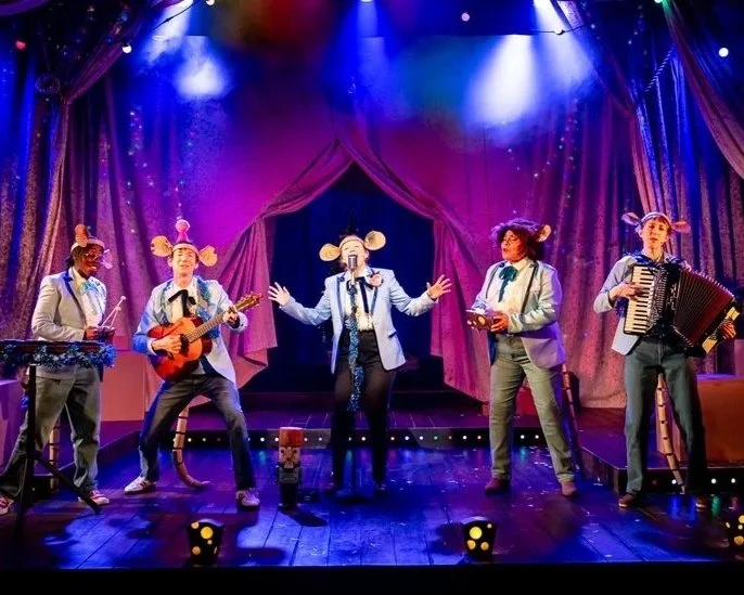 A group of performers dressed as musical mice sing and play instruments on a brightly lit stage, with sparkling curtains and colourful lighting creating a festive, theatrical scene.