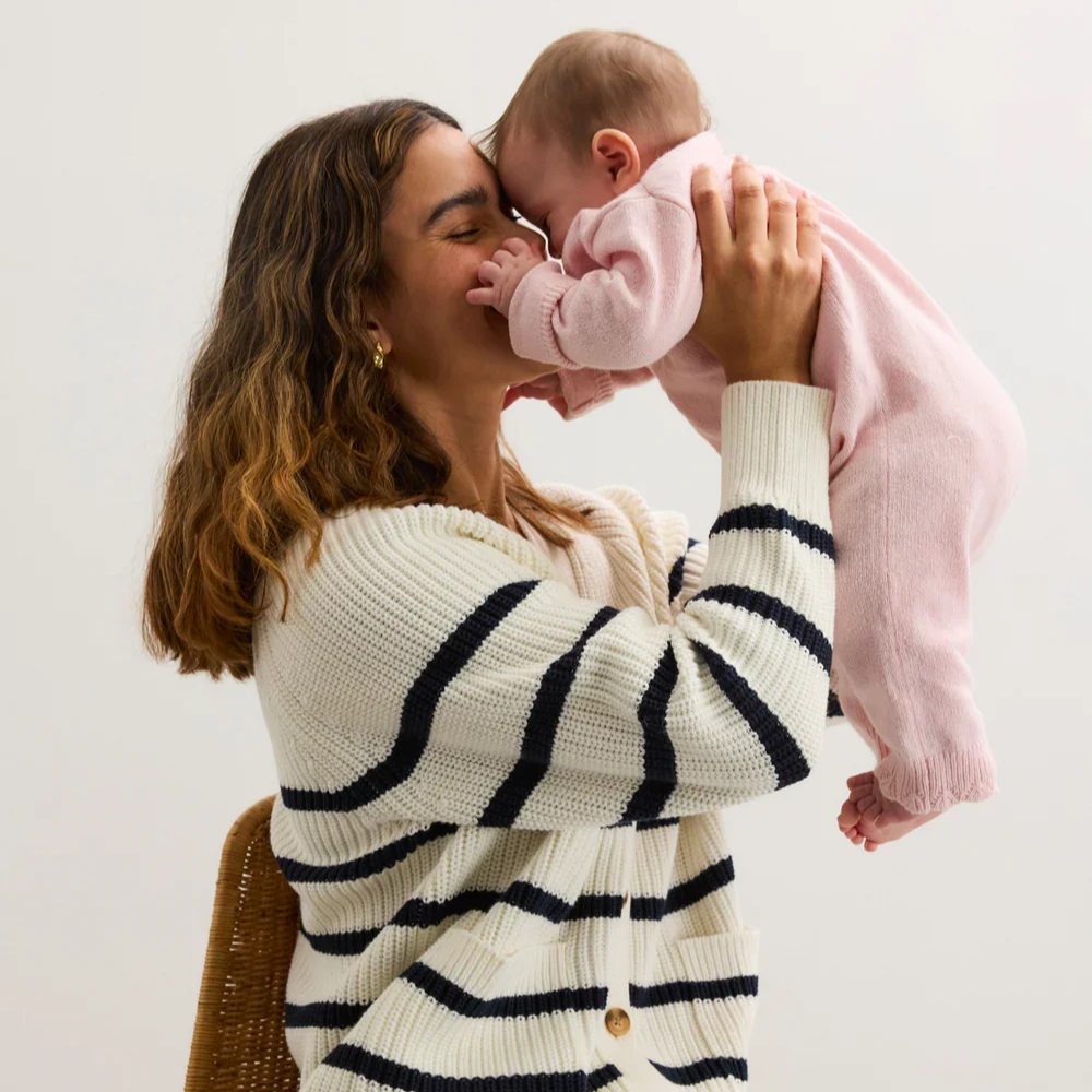 A mother holds her baby close while wearing the Hatch Cotton Marlow Stripe Cardigan, a soft cream knit with navy stripes and wooden buttons, styled with denim for relaxed maternity and postpartum comfort.