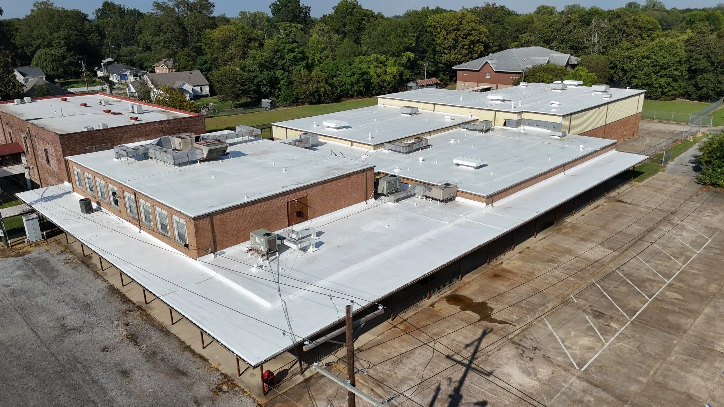 Aerial view of a flat rooftop with multiple buildings, HVAC units, and surrounding parking lot with empty spaces, with trees and neighborhood houses in the background.