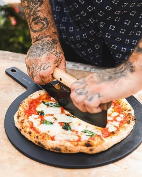 Person slicing a Margherita pizza on a black round cutting board with a large chef's knife, tattooed hands, outdoors setting.