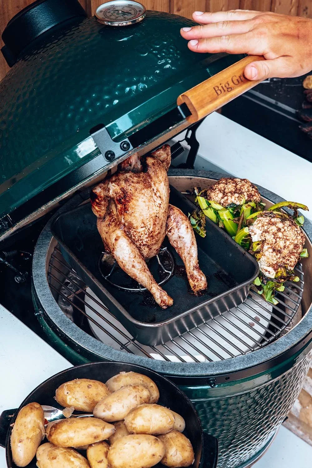 A person lifting the lid of a Big Green Egg grill to reveal a whole roasted chicken inside, with roasted cauliflower and greens on the side. A bowl of potatoes is in the foreground.