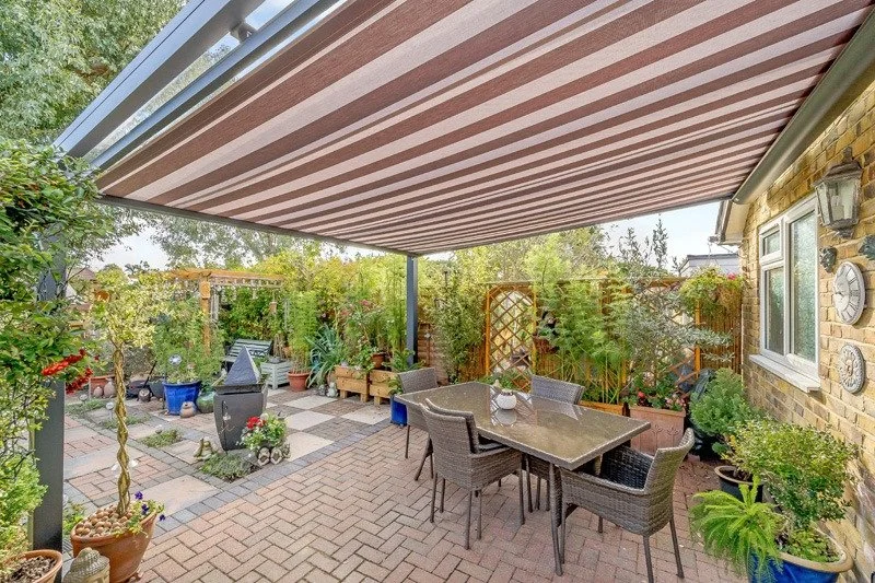Backyard patio with brick flooring, outdoor dining table with six chairs, lush greenery, potted plants, decorative trellises, and a retractable striped awning.