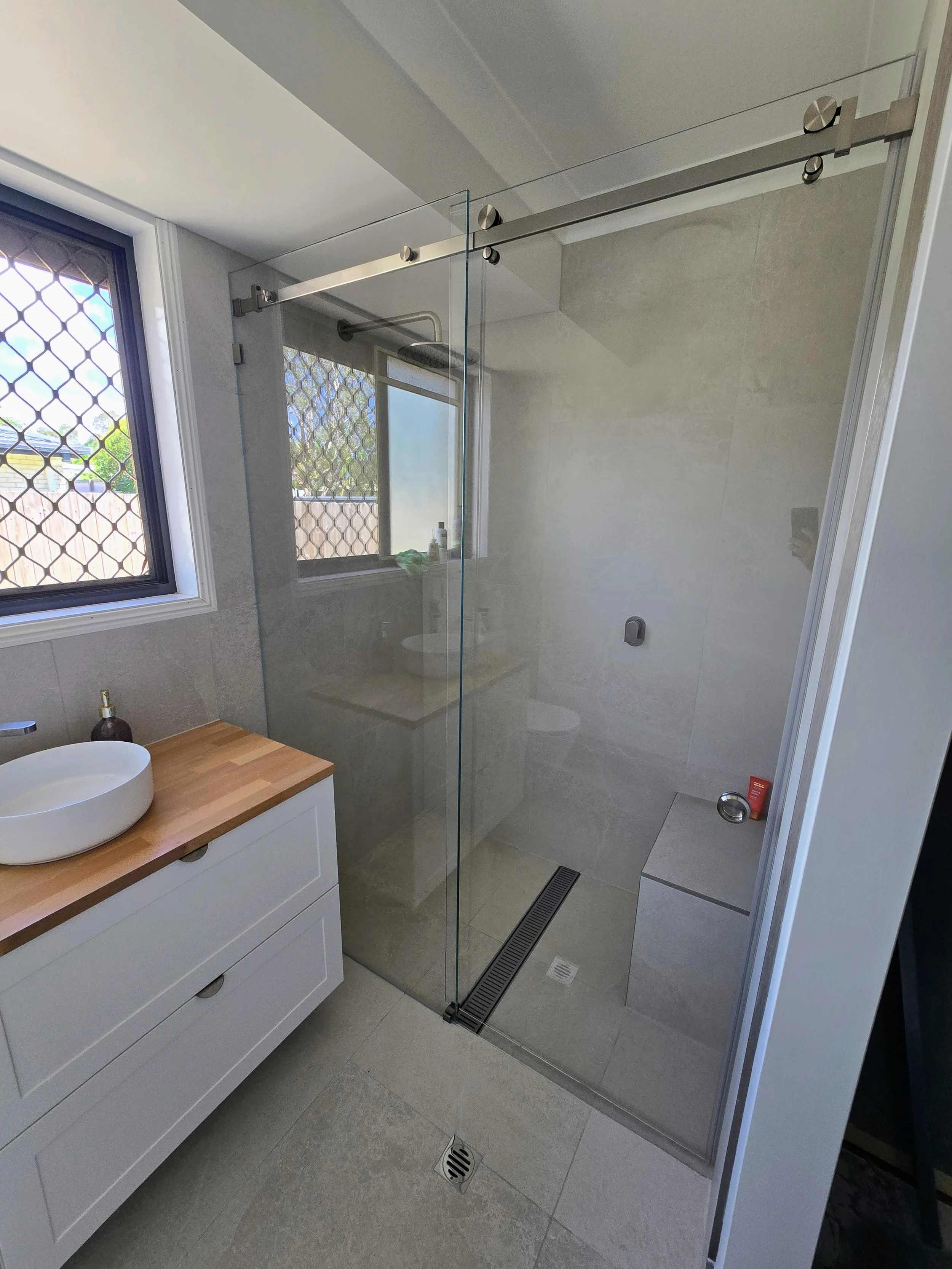 Modern bathroom with a vanity, sink, and a walk-in shower with a glass door. There is a window with a security screen, a drain in the floor, and a ventilation grille, with neutral-colored tiles on the walls and floor.