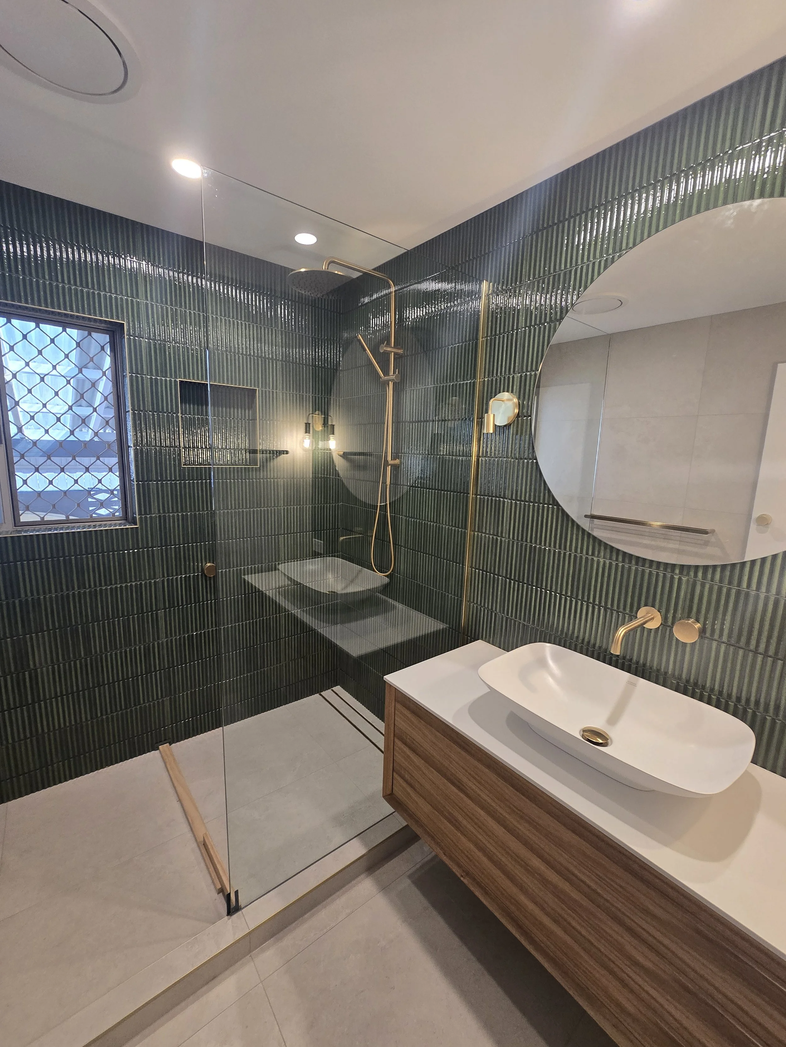 Modern bathroom with green tiled walls, a glass shower enclosure, a round mirror, a wooden vanity with a white sink, gold fixtures, a window with a grid pattern, and light-colored floor tiles.