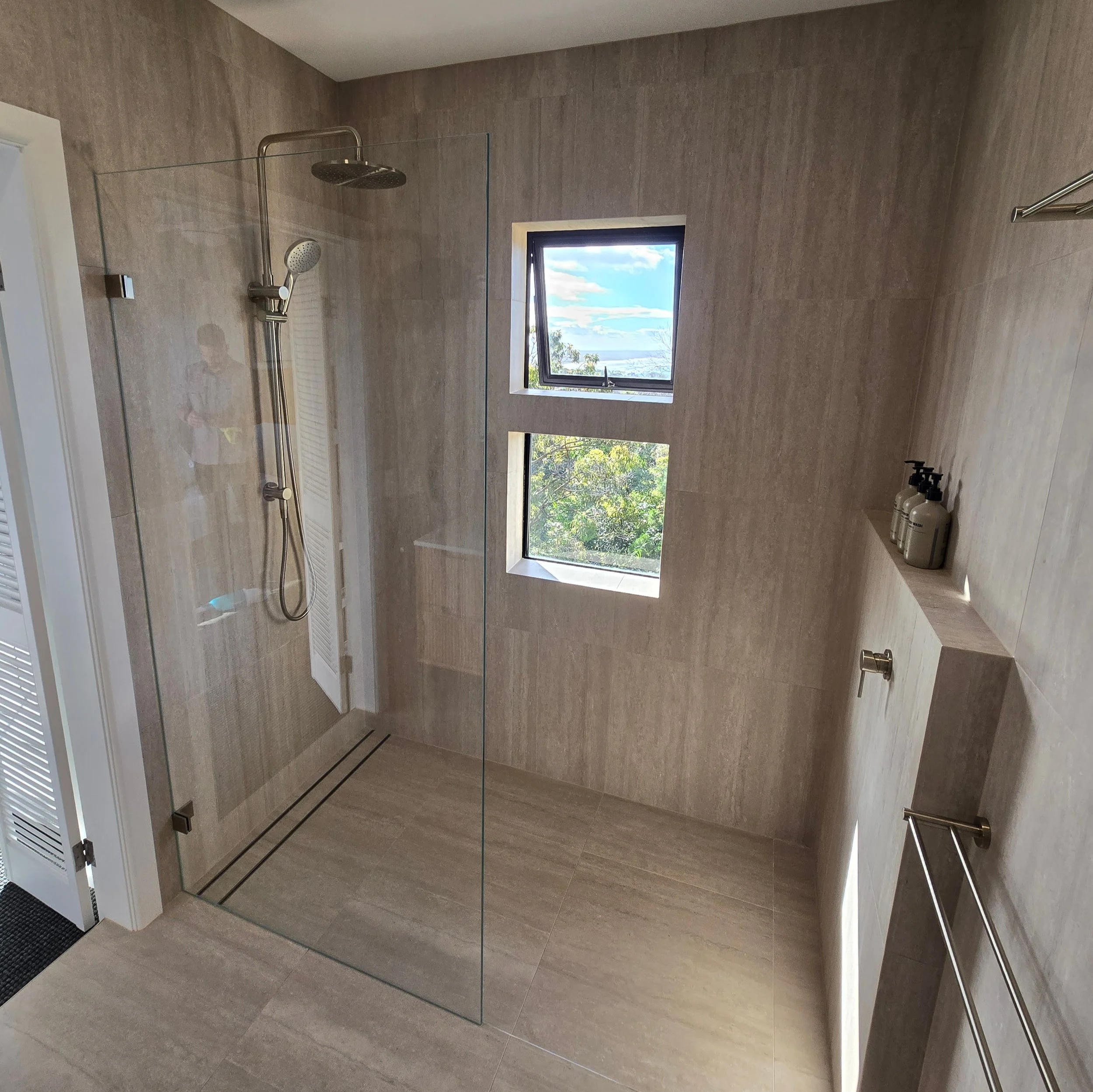 A modern bathroom with gray tiles on walls and floor, a glass shower enclosure, a rainfall showerhead, a window with a scenic outdoor view, and bottles of soap or shampoo on the sink.
